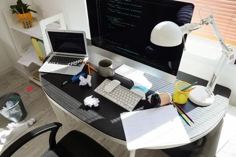 Messy programmers desk with laptop, computer monitor displaying code, and off 스톡 사진