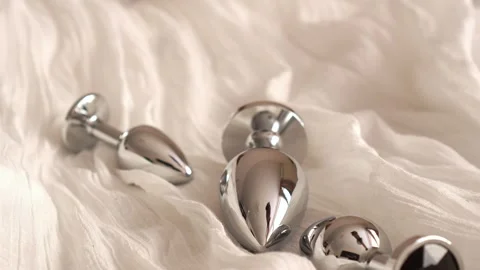 Metal butt plugs of various sizes with black rhinestones fall onto the bed. BDSM Stock Footage 319830202