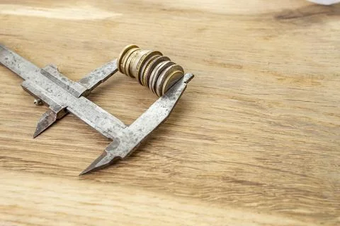 Metal caliper gripping a high stack of golden coins on a wooden desk, close u Stock Photos