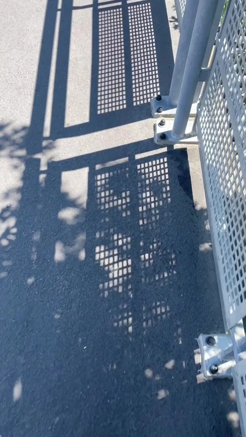 Metal fence casting shifting grid shadows across concrete walkway in bright sun Stock Footage 341661379