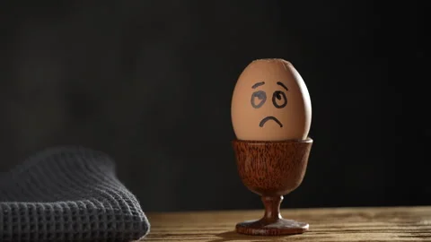 Metal spoon breaking boiled egg with sad face in egg cup on wooden table Stock Footage 304447975