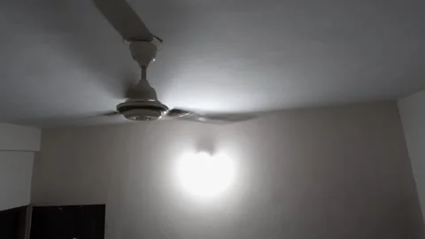 A metallic fan cuts through heavy, stagnant air in an underfunded living room at 動画素材 343784165