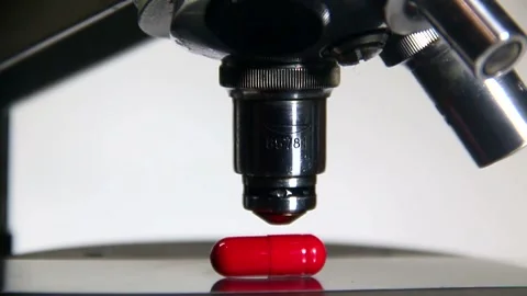 A microscope lens is lowered onto a red pill on a white background in macro Stock Footage 317310250