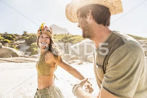 Mid Adult Couple Wearing Straw Hat And Feather Headdress Running On Beach, Cape