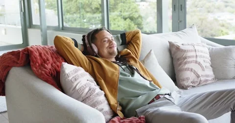 Mid adult man settling on sofa at home then lifting over-ear headphones sinking Stock Footage 341937344
