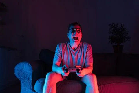 Mid adult man sitting on sofa and playing video games with controller. Neon Stock Photos