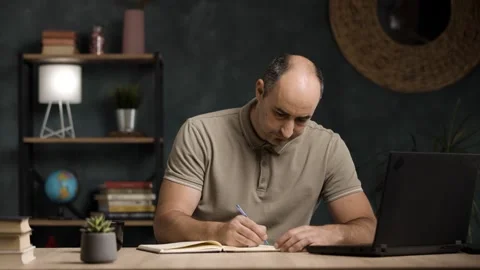 Mid Adult Man Taking Notes in Notebook While Working on Laptop at Home Stock Footage 342236868
