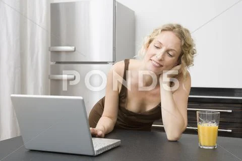 A mid adult woman leaning on a kitchen counter using a laptop Stock Photos