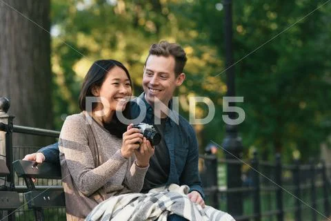 Mid Adult Woman Sitting On Park Bench With Boyfriend Taking Photographs On Slr