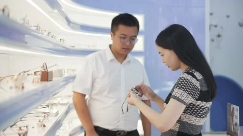 Mid-age consultant man in optical store helps to choose glasses for young woman Stock Footage 195140723