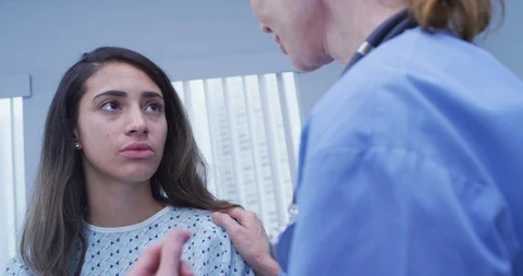 Mid aged nurse comforts young latina patient wearing hospital gown Stock-Footage 94457891