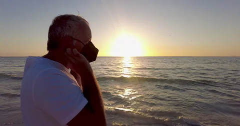 Middle age man by beach looking at golden sunset putting on a facemask. At ocean Video stock 218564059