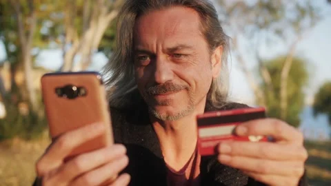 Middle age man customer holding credit card and smartphone sitting in the park. Stock Footage 223365147