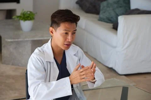 Middle aged Asian man in lab coat sitting at glass table in living room Stock Photos