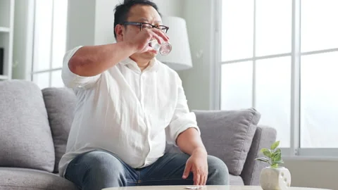 Middle-aged Asian man taking pill of medicine while sitting on sofa at home Stock Footage 232080925