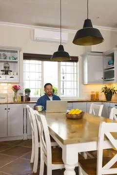 Middle-aged Asian man working on laptop at wooden dining table in kitchen, Stock Photos