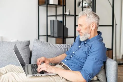 Middle-aged bearded man lying down on the couch with a laptop Stock Photos