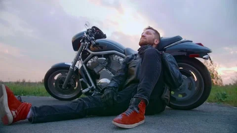 Middle-aged biker with beard sits by motorcycle at sunset Stock Footage 154815378