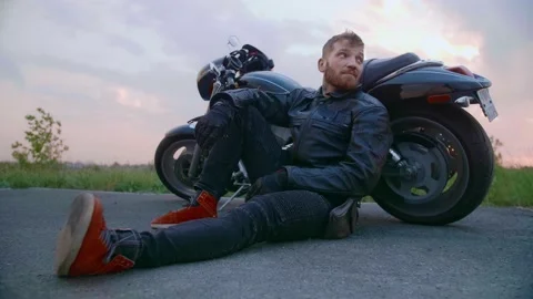 Middle-aged biker with beard sits by motorcycle at sunset Stock Footage 154915389