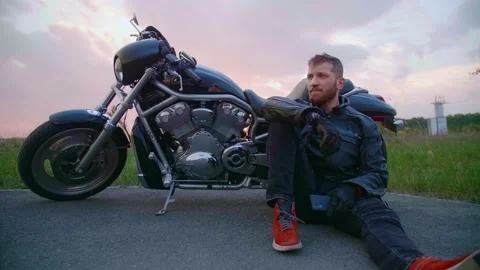 Middle-aged biker with beard sits by motorcycle at sunset Stock Footage 154915776