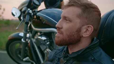 Middle-aged biker with beard sits by motorcycle at sunset Stock Footage 154916297
