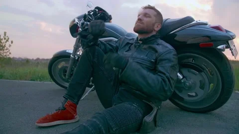 Middle-aged biker with beard sits by motorcycle at sunset Stock Footage 154916346