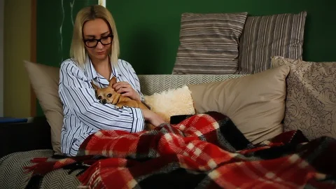 A middle-aged blonde is at home on the sofa under a blanket stroking a cute dog  Vidéo 114381022
