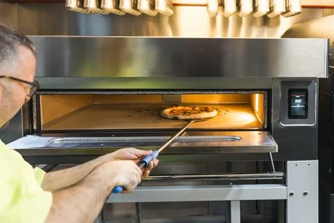 Middle-aged chef taking cooked pizza out of an electric oven, bakery kitchen Stock Photos