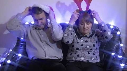 Middle-aged couple puting on santa hat and reindeer horns sitting on a couch Stock Footage 70698293