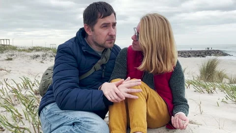 Middle-aged couple sitting on a beach, talking and cuddling affectionately Stock Footage 312230034