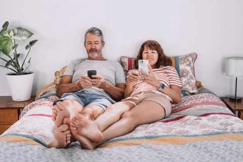 Middle-aged couple using their mobile phones laying on bed with their legs Stock Photos