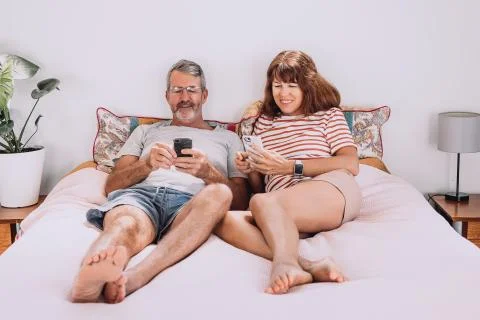Middle-aged couple using their mobile phones laying on bed with their legs Stock Photos