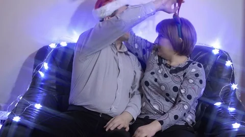 Middle-aged couple wearing santa hat and reindeer horns sitting on a couch and Stock Footage 70698308