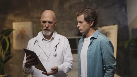 Middle-aged doctor explains diagnosis to male patient using tablet at check-up Video stock 342749782