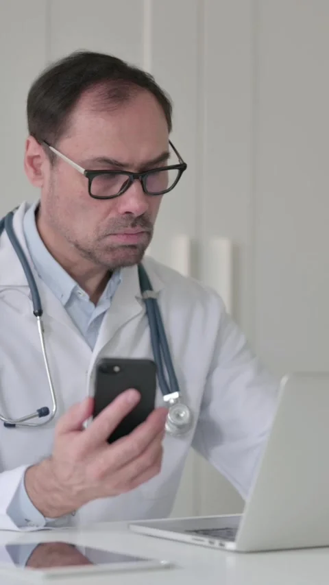 Middle Aged Doctor using Smartphone while Working on Laptop in Office, Vertical Vídeo Stock 339583546