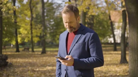 A middle-aged handsome Caucasian man works on a smartphone in a park in fall - Vídeo Stock 201775331