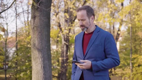 A middle-aged handsome Caucasian man works on a smartphone and walks in a park Video stock 201780481
