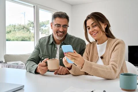 Middle aged happy couple using smartphone relaxing at living room table at home. Stock Photos