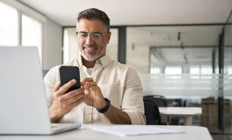 Middle aged Hispanic manager ceo businessman using cell phone mobile app, laptop Stock Photos