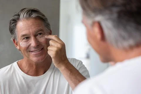 Middle-aged man applying under-eye patches at bathroom vanity mirror adjusting Stock Photos