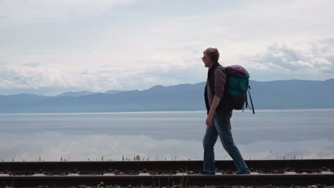 Middle aged man with backpack walks along rails of old railway along Lake Baikal Stock Footage 234049945
