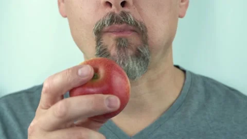 Middle aged man with beard holds in hand and eats heathy red apple, close up 4K Stock Footage 157115682
