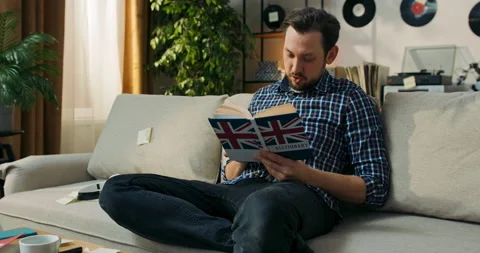 A middle-aged man with a beard is sitting on a couch with an English dictionary Stock Footage 200831677