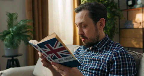 A middle-aged man with a beard is sitting on a couch with an English dictionary Stock Footage 200831920