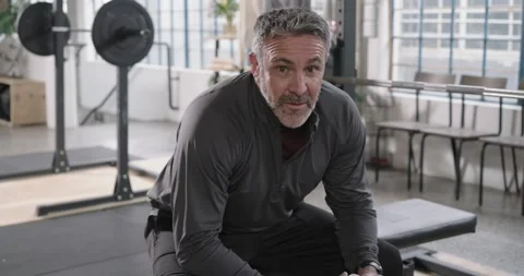 Middle-aged man in dark top sitting on bench in gym holding shaker, recovering Stock Footage 338338140