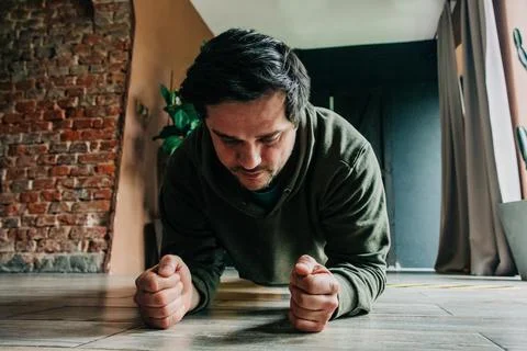 Middle-aged man doing plank and push-ups on floor near window in tracksuit,.. Stock Photos