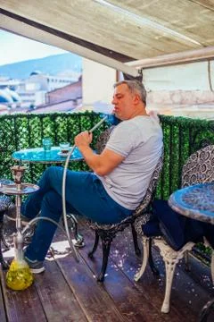 A middle-aged man dressed casually is smoking hookah (shisha) while sitting a Stock Photos