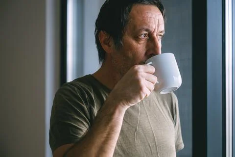 Middle aged man drinking coffee from a cup while standing by a window, thou.. Stock Photos