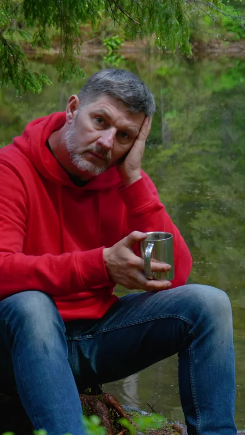 Middle aged man drinking hot beverage beside calm forest lake 库存影片 334390065