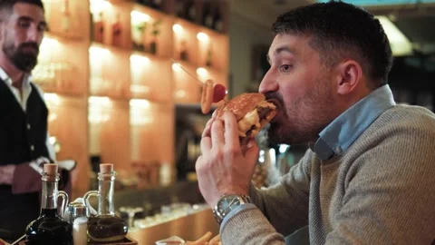 Middle-aged man eating fast food, enjoys eating hamburger in bar Stock Footage 171166734